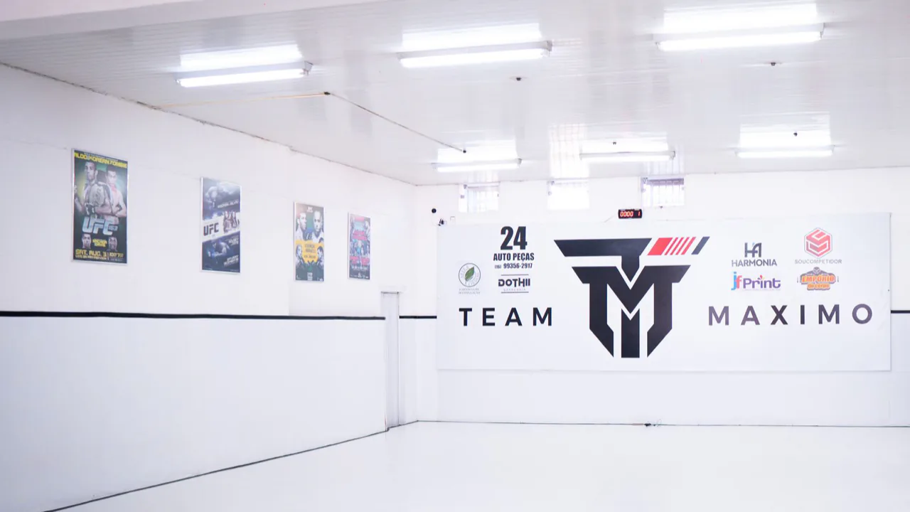 Tatame branco e amplo da academia, com a parede do logo Team Máximo ao fundo